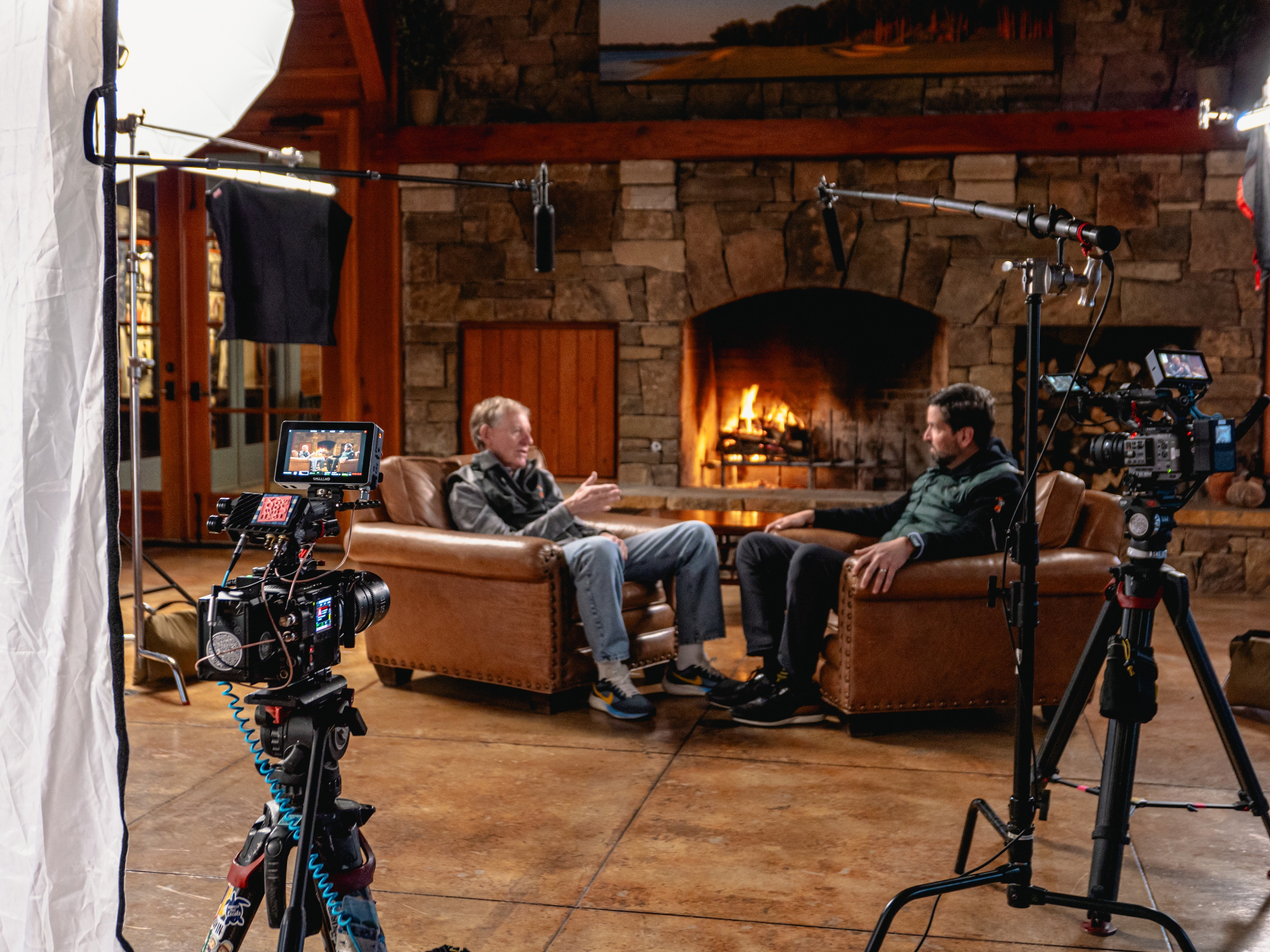 Behind the scenes — two subjects seated in leather armchairs in front of a stone fireplace, filmed by a documentary crew with cameras, lighting, and boom mics visible.
